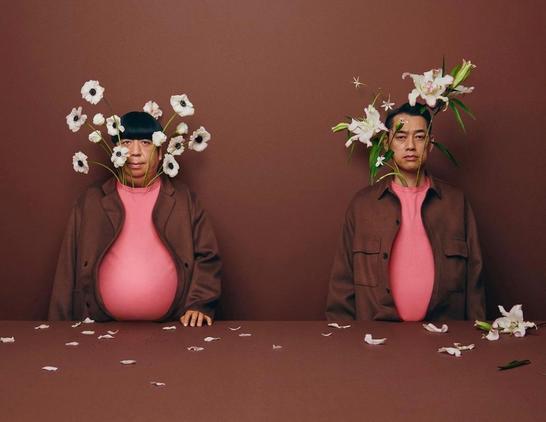 Twee mannen zitten aan tafel achter, ogenschijnlijk, een roze vaas met witte bloemen.  De mannen dragen bruine jasjes over roze t-shirts. De kleding is zo gedrapeerd en gevormd dat het bloemvazen lijken. In de shirts zijn witte bloemen gestoken. Zo lijkt het alsof er bloemen in een vaas staan.