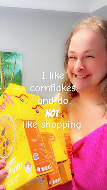 Me with a big cheesy grin on my face, barely holding five packs of Freedom Classic Corn Flakes (there were three more in my order 😁) in one hand, against myself. Caption says: I like cornflakes and do NOT like shopping.