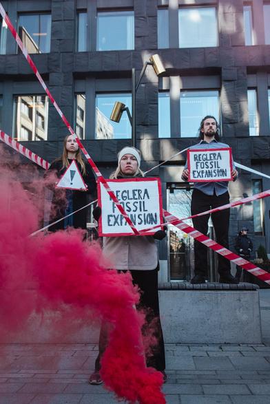 Folks standing with placards saying "Reglera fossils expansion" and red smoke coming out from below, we can also see the emergency tape in between 3 people in the photo.