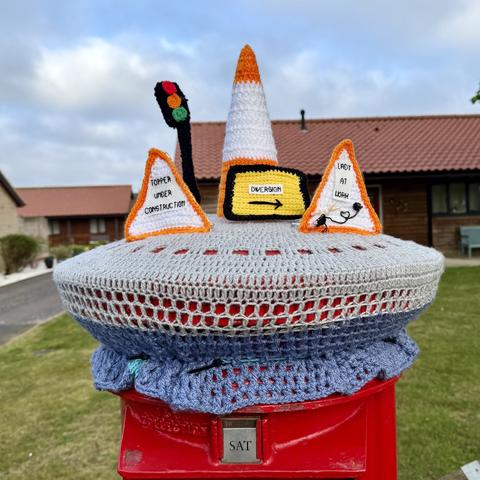 Red post box with a knitted topper. It’s an orange and white cone with triangle signs next to it saying TOPPER UNDER CONSTRUCTION, DIVERSION, and LADY AT WORK. The latter has a tiny set of knitting needles and yarn next to it. There’s also a traffic light behind all these. 

It’s beautiful.