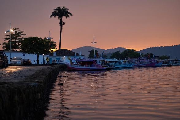 On the left side the quay wall of the port of Paraty is shown, with the sea reflecting the orange and blue sky in the middle and to the right. In the middle of the photo colorful fisher boats are seen in front of mountains covered with Atlantic rainforest before the orange-blue morning sky. On the left the street lights are still on and a person with an umbrella is cycling.