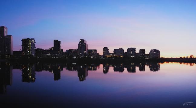 Das Foto zeigt die Skyline der Seestadt Aspern mit mehreren Hochhäusern und Wohngebäuden, die sich im ruhigen Wasser des Asperner Sees spiegeln. Der Himmel ist in sanften Farbtönen von Blau, Lila und Rosa gefärbt - Abenddämmerung. Einige Fenster der Gebäude leuchten, während der Rest der Stadt bereits im Schatten liegt. Die Spiegelung im Wasser verstärkt die ruhige und harmonische Atmosphäre, während der Horizont einen sanften Farbverlauf zeigt, der in warmen Orangetönen ausklingt.