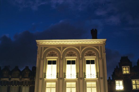 The upper part of a neo-classical facade at night, with other old facades (in different styles) on its left and right. The tall windows shine a bright light. Behind the buildings, filling almost half of the picture, is a dark blue sky with dark grey clouds.