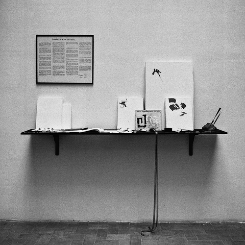 Une étagère sur un mur nu, sur laquelle repose divers publications et document. Au mur, encadré un manifeste lettriste. Photo noir et blanc.