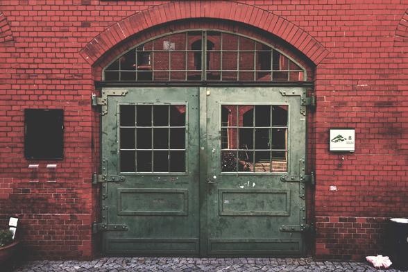 Eine grüne, doppelflügelige Metalltür mit Fensteriensätzen und einem fast halbkreisförmigen Fensterbogen in einer roten Backsteinwand