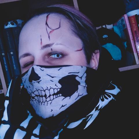 Alternative guy with skeleton mask (no affiliation to any stupid groups), skeleton hoodie and skeleton gloves, with a bit of artistic makeup, white lenses, one eye winking, undercut, pink top hair, showing the so-called "devil horns" sign. Book shelf behind him, v with some books and misc items, such as black and cyan squishmallow bat. Image is dark as per suggested vibe.