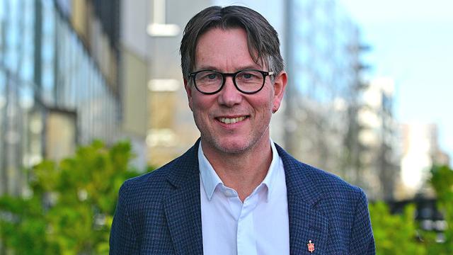 Porträt von Knut Refsdal. Ein ein lächelnder Mann mittleren Alters mit braunem Haar und Brille. Er trägt einen dunkelblauen, gemusterten Blazer über einem weißen Hemd. Der Hintergrund ist unscharf und zeigt eine moderne Architektur und etwas Grün.