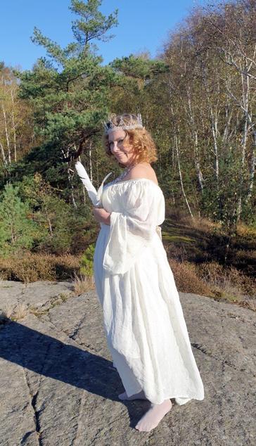A white middle aged woman with curly strawberry blonde hair wearing a white, loose dress, and  crown of silver coloured wire and clear white beads and prisms. She is holding a white feather in her hand. She is standing on bedrock, and behind her are trees.