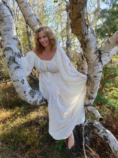 A white middle aged woman with curly strawberry blonde hair wearing a white, loose dress, sitting on a branch in a birch tree.