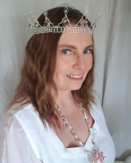 The face and upper torso of a white middle aged woman with brown, wavy hair, wearing a white, dress, and  crown of silver coloured wire and clear white beads and prisms.