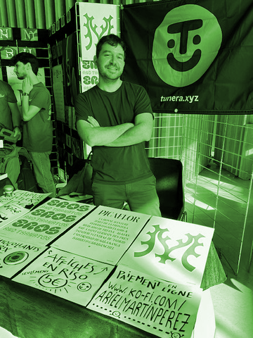 Monochrome green tint photograph of a middle aged man smiling at the camera and crossing his arms, he's surrounded by a lot of typographic postera and catalogs including a big flag with a logotype that reads tumera.xyz
