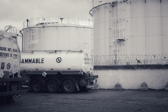 Un camion-citerne marqué "FLAMMABLE" est stationné devant des réservoirs de stockage de carburant blancs, entourés d'une clôture en fil barbelé. Les réservoirs sont équipés d'échelles et de plateformes pour l'accès. L'environnement est industriel et nuageux.