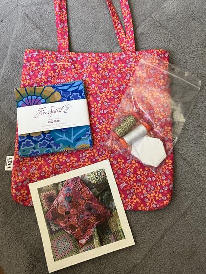 Een Leonieke-gemaakt tasje met rode bloemetjespatroon, een pakje kleine lapjes met blauwe en paarse patronen (voor de kenners: een “charm pack”), een boekje met patroon met een foto van een kussensloop voorop, en een zakje met zeskantige papieren malletjes en twee klosjes garen