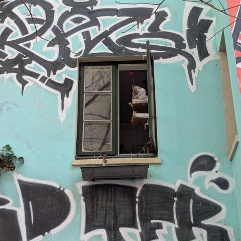 Fenster in einer mit Graffiti versehenen Außenwand eines Gebäudes im Hamburger Gängeviertel. Hinter dem geöffneten Fenster hängt ein künstlerisches Gebilde.
