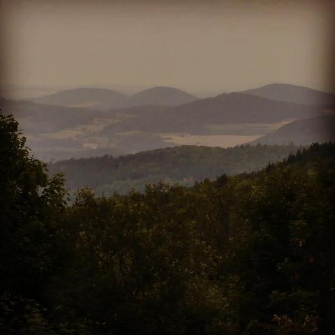Das Bild zeigt eine malerische Landschaft mit einer Reihe von sanften Hügeln, die sich in der Ferne im Dunst verlieren. Im Vordergrund sind dichte Wälder zu sehen. Die Farben sind gedämpft und abgedunkelt, so dass die Szene ruhig, aber auch etwas beklemmend wirkt.