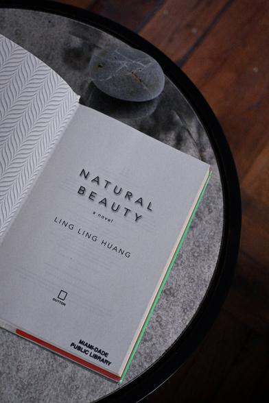 Book, Natural Beauty by Ling Ling Huang, set on mirrored table. Adorned with two gray stones. Book opened to title page. Miami Public Library stamp.
