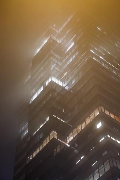 Tall office building at night, seen from below, windows illuminated, casting hazy glowing into the fog surrounding it.