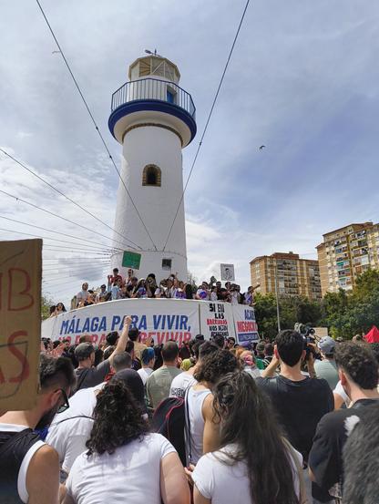 Una gran multitud de manifestantes se congrega en una plaza o parque, abarrotada de gente que sostiene pancartas y carteles. La mayoría de los manifestantes parecen ser adultos, y muchos muestran su apoyo con gestos activos como levantar los brazos. Una pancarta grande y prominente en el centro de la imagen proclama "Vivienda por Derecho". Se puede observar equipo de sonido en el suelo, sugiriendo un evento organizado. El ambiente general es el de una protesta o manifestación pacífica, con una gran cantidad de personas que participan en la demostración.  El fondo muestra árboles verdes y parte de un edificio, indicando una ubicación al aire libre en una zona urbana.