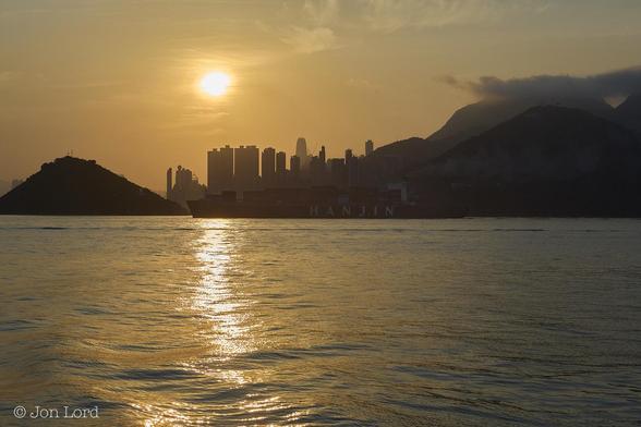 This Is A Colour Sunrise Seascape / Cityscape Photo In Landscape Format. The Lower Half Of The Image Is Filled With An Expanse Of Calm Water, Mostly A Golden Colour From The Reflection Of The Rising Sun. There Is A Bright Streak Of Light Stretching Along The Water From Under The Sun Towards The Camera. In The Middle Ground And The Centre Of The Photo Is A Large Container Ship Under Way From Right To Left. The Vessel Is Deep In Shadow Though The Name Of The Shipping Line 'HANJIN' Is Clearly Visible In Large White Letters On The Ships Black Hull. The Stacked Shipping Containers Can Be Made Out Forward Of The White Accommodation / Wheelhouse Superstructure. There Is A Trail Of Grey Smoke Trailing About One Ships Length Above And Aft Of The Ship. On The Far Side Of The Ship Is A Number Of Large Hills, Again Deep In Shadow, Some With Wisps Of Cloud Covering The Peaks. In The Background Is The Silhouette Of Numerous Skyscrapers With The Bright Orb Of The Still Low Sun A Short Distance Above. The Sky Is Nearly Clear With Only A Small Amount Of Near Transparent Cirrus High Cloud Present. 
The Location Is The Busy East Lamma Shipping Channel. The Terrain And Skyscrapers Are Hong Kong Island (2015).
