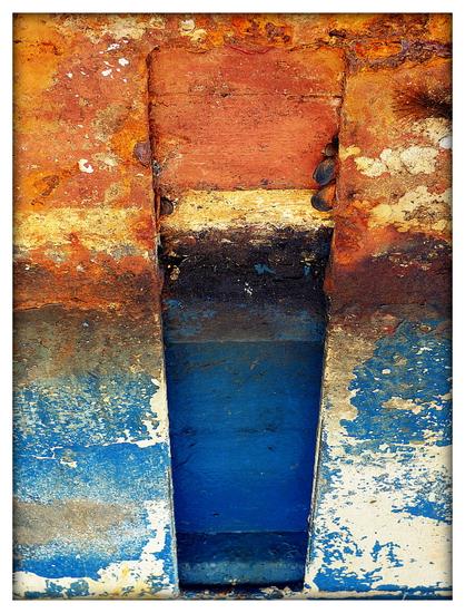 A colour photograph showing a smal section of boat hull under refit. The composition transitions vertically from rust to blue with a worn strip of white separating. A darker monolithic shape passes through the centre of the frame, darkened by being recessed.