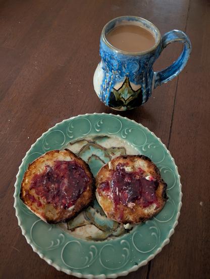 A cup of coffee and an english muffin with butter and red raspberry preserves.