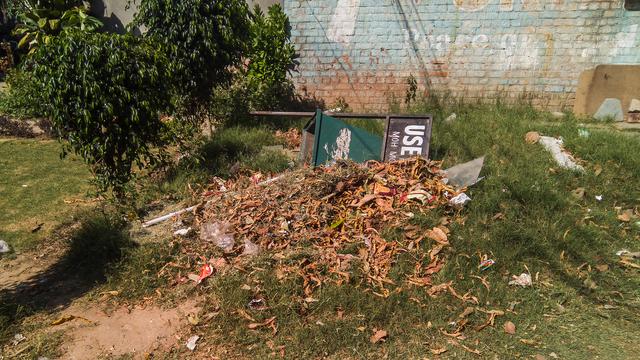 A photo of a trashcan laying sideways in a pile of collected browned leaves. The trashcan is green in colour and is made of metal, with a label on top which reads "USE ME" presumably. To the left of the trash,there are two bushes/hedges with green leaves. In he background, there is a brick wall with fading blue colour painted on it for some sort of an advertisement. There is also green grass surrounding the trash can and leaves on the ground.