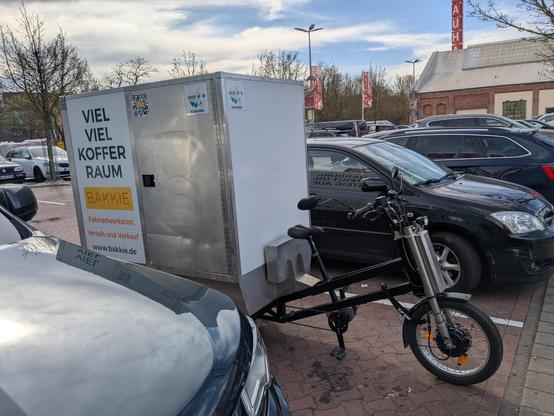 Foto eines E-Lastenrads mit geschlossenem Aufbau auf der Ladefläche, aufgenommen auf einem Baumarkt-Parkplatz.