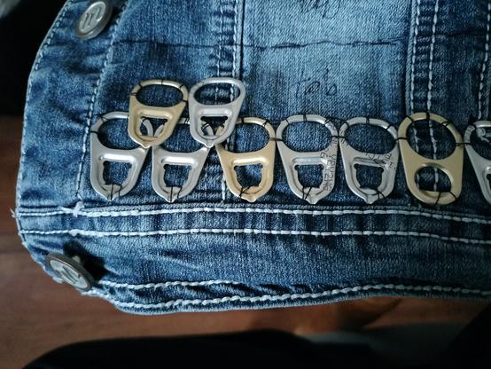 The corner of a denim jacket, on the bottom edge next to the buttons. Right above the waistband, there's a row of cat food can tabs sewn into the denim. They're in a pattern of 2 silver-colored can tabs, 1 gold-colored can tab. I've also started the next row, with the first gold tab on top of two silver tabs from the first row. In the first row, the part of the tabs where all human puts our finger to open the can will be covered by the other side of the tabs in the second row.
Future rows of can tabs are sketched out on the jacket in sharpie above the current rows. Each row is a big rectangle with the word "tab" in it.