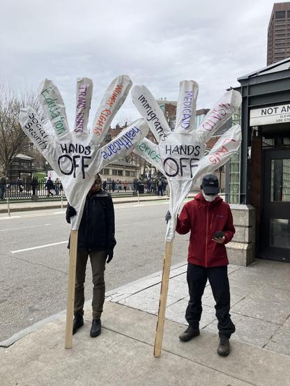 Two protesters holding giant white hands on poles, with the text "hands off" on the palms, and education, trans rights, immigrants, medicare, women's rights and more on the fingers.