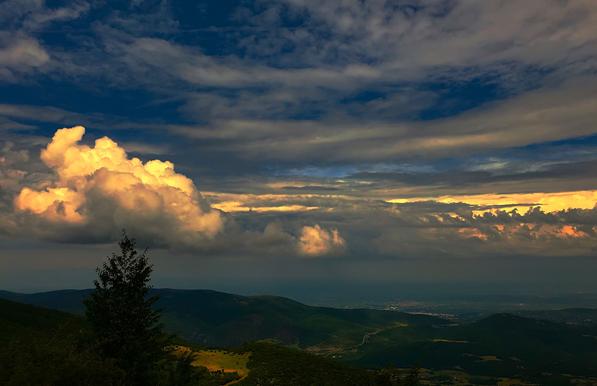 A scenic view , uphill a mountain looking from above , sky full of clouds with the sun giving those reddish colors upon them.