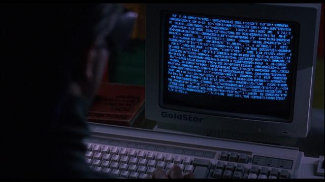 A frame from the 1992 movie, "Sneakers", shows a computer screen willed with structured but incomprehensible white text on a black background. The light from the GoldStar CRT monitor illuminates a keyboard in front and a dark blurry person in front of it in a dark room.