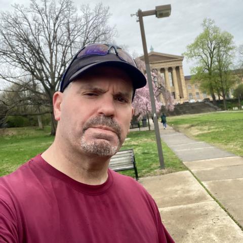 A man in a maroon shirt and black cap takes a selfie in a park with trees and a building in the background. The sky is cloudy, and there is a path and a bench nearby.