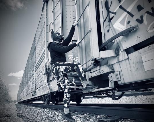 A dramatic monochrome scene captured in cool blue tones showing a figure in a sleek black neoprene puppy hood scaling the side of a massive freight train. upward with bare paws, demonstrating athleticism. Their body is clothed in a fitted black top and eye-catching black and white camouflage pants that contrast sharply against the industrial metal backdrop. The freight car's riveted steel surface stretches into the distance, marked with various industrial warnings. The composition creates a powerful sense of adventure and rebellion, with the figure silhouetted against the vast train that extends along tracks of scattered gravel. The moody blue-tinted filter adds an atmospheric, almost cinematic quality to this urban exploration moment. 

#PupPlay #TrainHopping #UrbanExploration #KinkLife #IndustrialAesthetic #MonochromePhotography