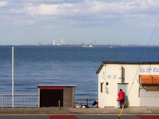 This image shows a coastal scene with a small building in the foreground, likely a shop, as indicated by the Japanese writing on it. There's a person in a red jacket standing beside the building. The background features a large body of water, probably a sea or a bay, with several visible boats and a distant skyline under a cloudy sky.