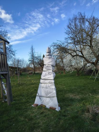 In einem Garten steht eine pyramidenförmige Konstruktion, die mit einem weißen Vlies umwickelt ist.