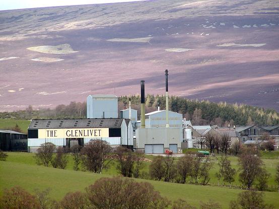 Glenlivet Distillery in Moray. The image shows a distant view of a distillery with the name The Glenlivet in large capital letters on its side. There are rolling fields in the foreground and trees and then rising moorland in the background with the skyline partly visible at the top of the frame.