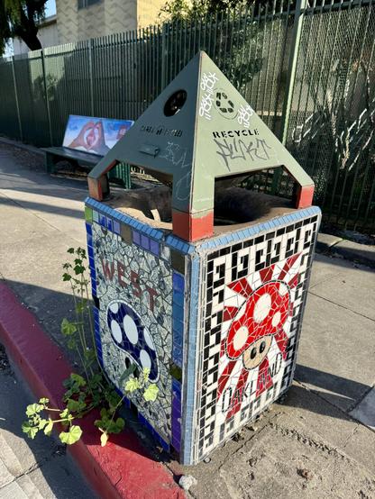 A trash can with unique mosaic art on the sides. The can is viewed at an angle, so you can see two sides of it. One side has a mosaic of a blue mushroom and text saying "West". The other side has a red-topped mushroom and text that says "Oakland". The mushrooms are the same as the 1-up mushrooms in the Super Mario games.