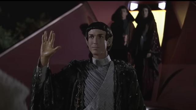 A Vulcan (Cully Fredricksen) stands at the bottom of the ramp in front of the Vulcan ship, the T'Plana-Hath making a hand gesture with the palm forward, thumb extended, and middle and ring fingers parted in the traditional Vulcan salute. Two Vulcans stand behind him near the top of the ramp at the entrance to the ship.