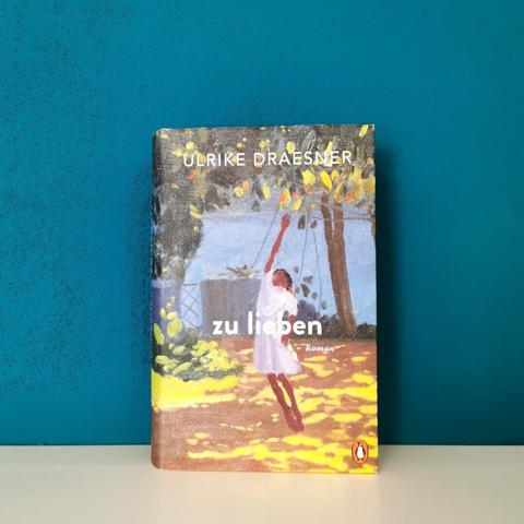 Auf dem Bild ist das Buch "zu lieben" von Ulrike Draesner fotografiert. Es steht auf einer glatten, weißen Fläche an eine dunkelgrüne Wand gelehnt.