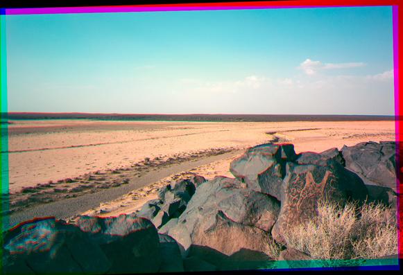 A view of a sandy desert mud flat, with basalt boulders in the foreground. There is a small camel petroglyph in the bottom right.
