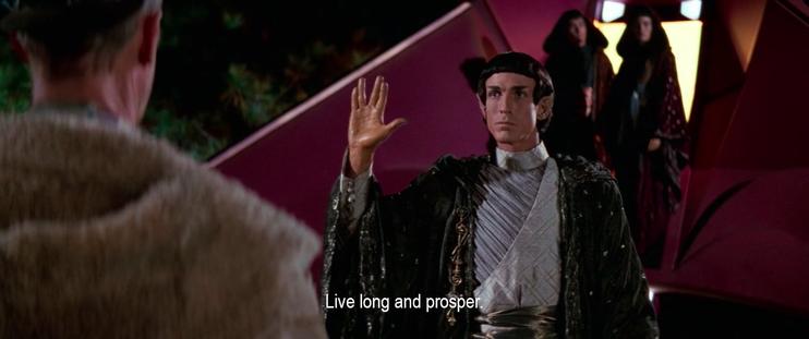 A Vulcan has emerged from the ship and lowered his hood, revealing his pointed ears. He holds his hand up in a gesture of greeting, saying, “Live long and prosper.”