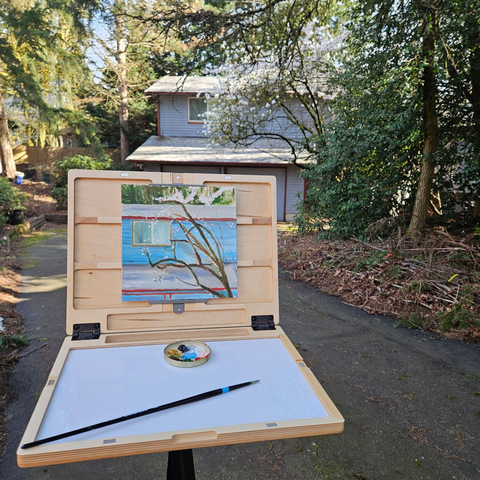 A painting outdoors of a blue house with red trim and a pacific dogwood and cherry tree blooming in front of it. Photo shows both painting on the easel and house