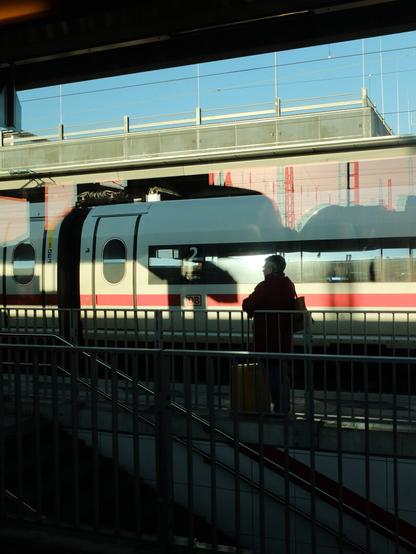 A person in silhouette wearing a red coat stands on a train platform, waiting beside a white and red Deutsche Bahn (DB) high-speed train. The scene is backlit, creating a dramatic contrast with the bright blue sky visible through the station's overhead walkway. Platform number 2 is visible on the train. Metal railings and stairs can be seen in the foreground, with the station architecture framing the composition.