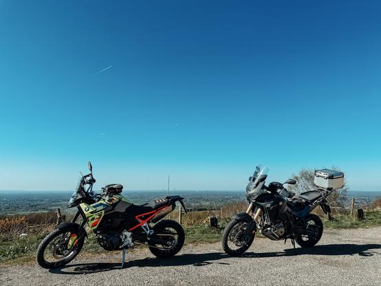 Two motorcycles parked in front of a view of Cheshire. The left hand bike is a BMW F900GS adventure bike in black/yellow with a red subframe, and the right hand bike is a Honda Africa Twin CRF1100L adventure bike in black. The sky is blue with not a cloud in sight.