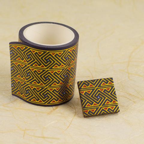A small square (25mm and about 3mm thick) is carefully wrapped in a bright printed Celtic key or maze pattern in purple, red, and metallic gold. It sits on one edge next to a roll of washi tape with the same pattern against a background of off-white fibrous handmade paper.