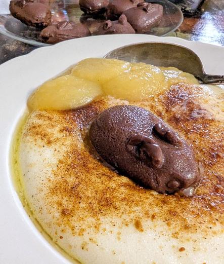 Tiefer, weißer Teller mit Grießbrei, braunen Gewürzen, einem Rand aus flüssiger Butter und einigen Esslöffeln Apfelmus an der Seite. In der Mitte sitzt ein hasenförmiges kleines Gebilde aus Schokopudding, ein Esslöffel steckt im Brei. Im Hintergrund ein gläserner Teller mit weiteren Bunnys.