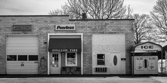 The front of an older building housing a tire store.  To the right is an ice machine covered by a metal canopy that says "ICE" on it.
