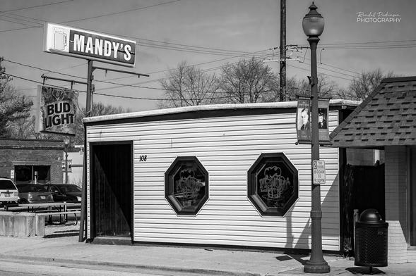 A small white sided 1 story building with 1 door and two octogon shaped windows.  A sign has a picture of a beer mug and the work Mandy's.  There is also a Bud Light sign.