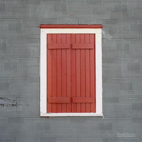 A white framed window in a grey concrete block wall has 2 red shutters that are shut and a matching red trim piece above the window.