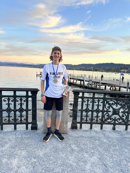 Ein junger Athlet (Liam) steht am Wasser, lächelt und hält eine Trophäe in der Hand. Er trägt ein "FINISHER 2025"-Shirt und Shorts. Hintergrund bietet einen malerischen Blick auf den See und die Berge unter einem bunten Himmel.